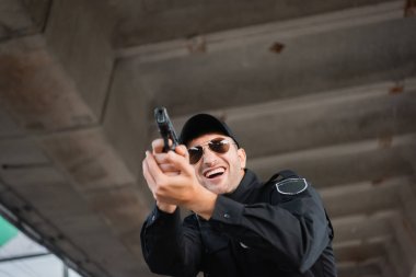 Güneş gözlüklü neşeli polisin ön planda bulanık bir şekilde silah tutmasının düşük açılı görüntüsü. 