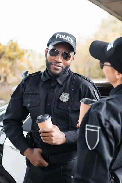 Sonriente Afroamericano Policía Con Taza Papel Mirando Cámara Cerca Colega —  Fotos de Stock