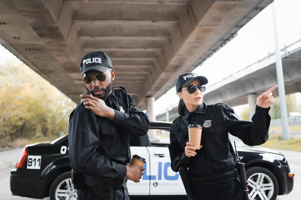 一个严肃的女警察用手指指指接近非洲的美国警察在室外模糊的背景下对着收音机说话 — 图库照片