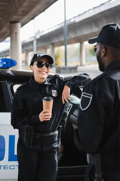 快乐的女警察拿着纸杯看着非洲裔美国同事 在模糊的背景下靠在巡逻车上 — 图库照片