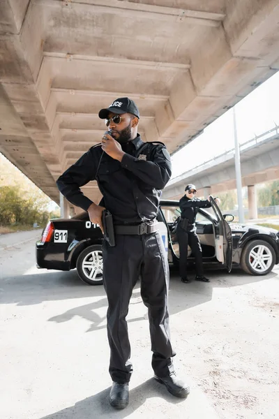 同僚とパトロールカーの近くの無線で銃の話をしているアフリカ系アメリカ人警官の長さ屋外でぼやけた背景に — ストック写真