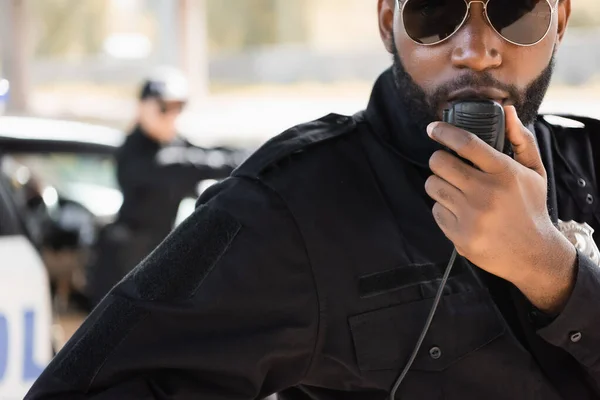 close up view of african american police officer talking on radio set with blurred colleague on background outdoors