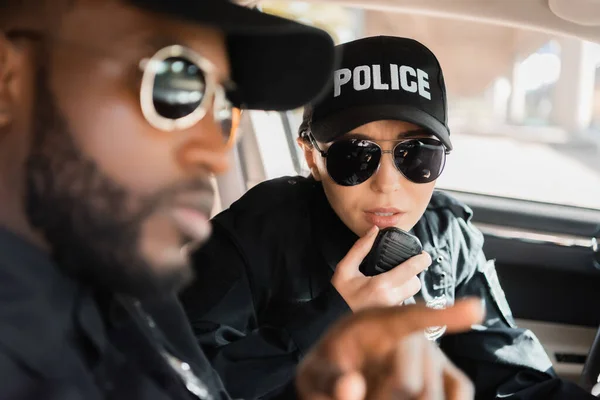 policewoman talking on radio set with blurred african american colleague on foreground in patrol car