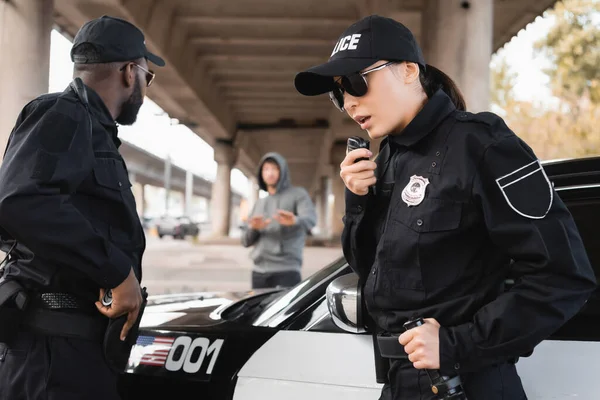 policewoman talking on radio set near african american colleague with blurred offender on background on urban street