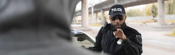 stock image serious african american police officer pointing with finger at blurred hooded offender on foreground on urban street, banner