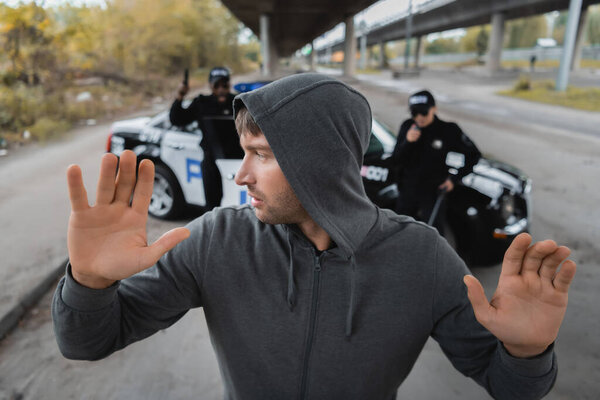 hooded offender with raised hands looking away with blurred multicultural police officers on background on urban street