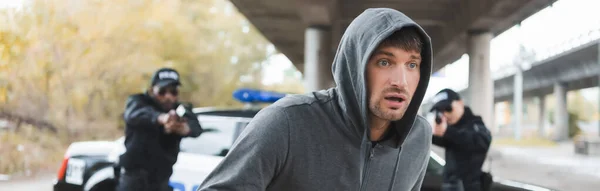 stock image scared hooded offender running from multicultural police officers on blurred background outdoors, banner