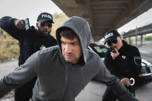 stock image dissatisfied hooded offender running from multicultural police officers on blurred background on urban street