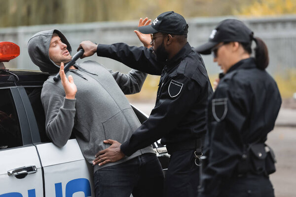 african american policeman with truncheon frisking offender with raised hands on blurred foreground outdoors