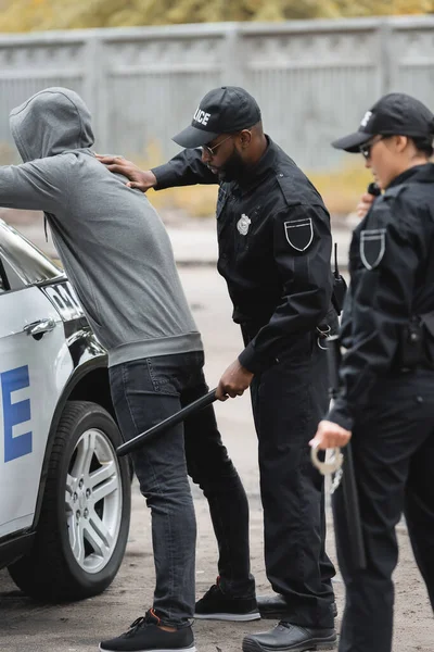 Африканский Американский Полицейский Дубинкой Обыскивающий Преступника Капюшоне Возле Патрульной Машины — стоковое фото