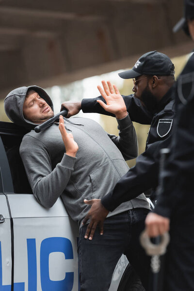 Африканский американский полицейский с дубинкой обыскивающий преступника в капюшоне с поднятыми руками на размытом переднем плане