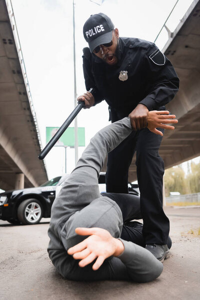 low angle view of aggressive police officer with truncheon arresting hooded offender lying on urban street 