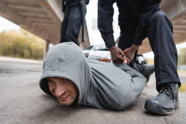 african american policeman handcuffing offender lying on street on blurred background outdoors