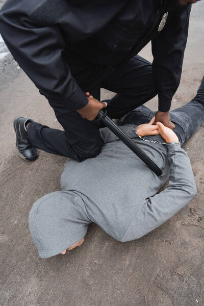 high angle view of african american policeman with truncheon frisking hooded offender lying on street