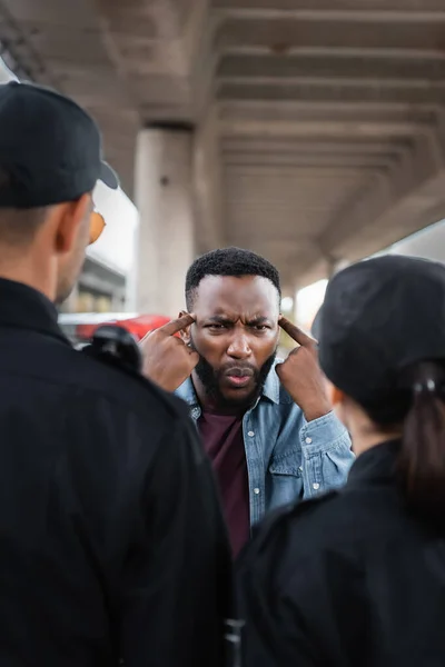 angry african american victim showing you are insane gesture while arguing with police officers on blurred foreground outdoors