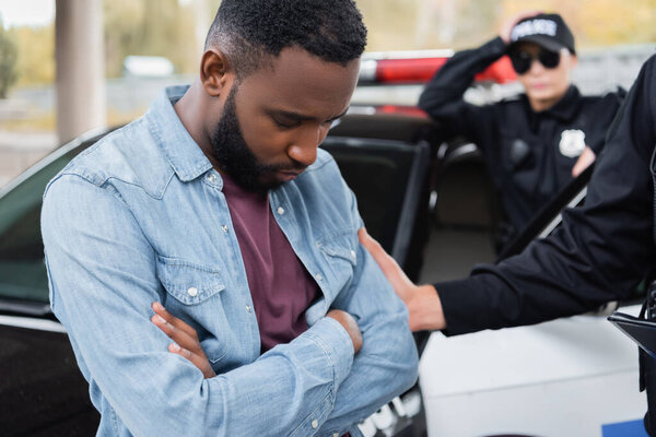 Sad african american victim standing near police officer and car outdoors 