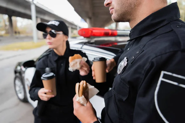 在城市街道上 警察拿着汉堡包和咖啡在模糊的背景下走近同事和汽车 — 图库照片