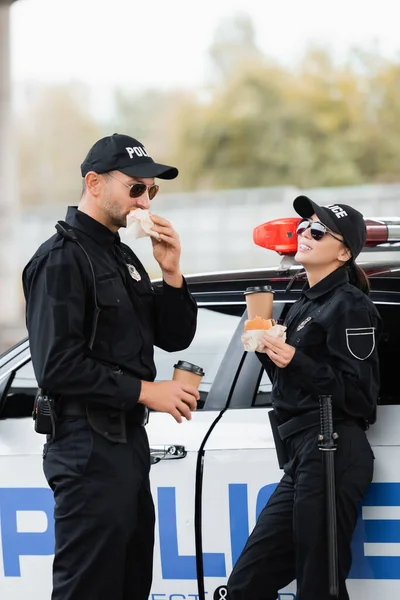 戴着太阳镜 拿着汉堡包和咖啡的女警察笑着走近同事吃饭和外面开车 — 图库照片