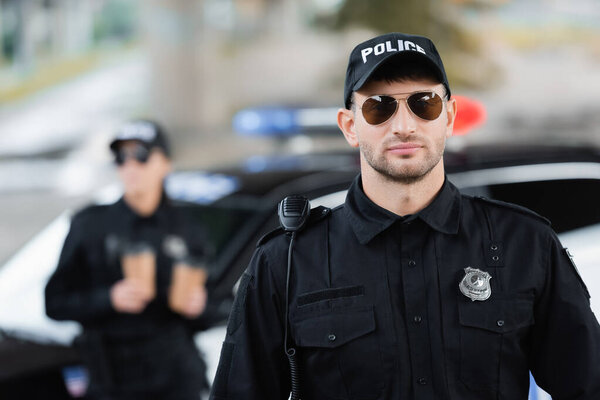 Officer of police in sunglasses near colleague and car on blurred background outdoors 