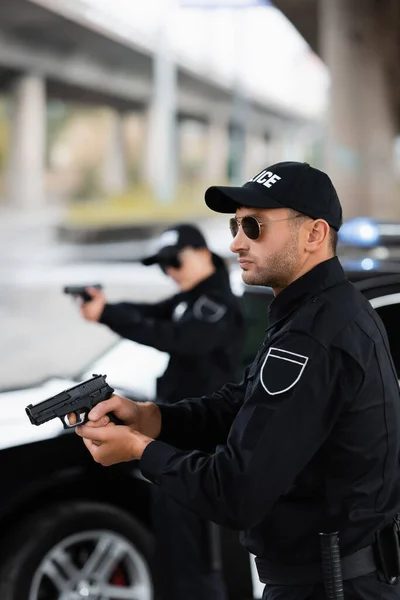 Polisman Solglasögon Med Skjutvapen Nära Kollega Och Bil Suddig Bakgrund — Stockfoto