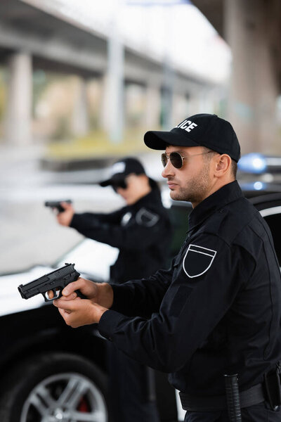 Police officer in sunglasses holding firearm near colleague and car on blurred background outdoors 