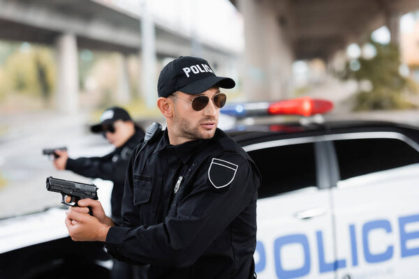 Policeman in sunglasses holding gun and looking away near colleague and car on blurred background 