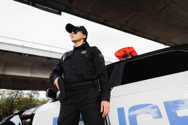Policewoman in sunglasses holding gun in holster near car on urban street