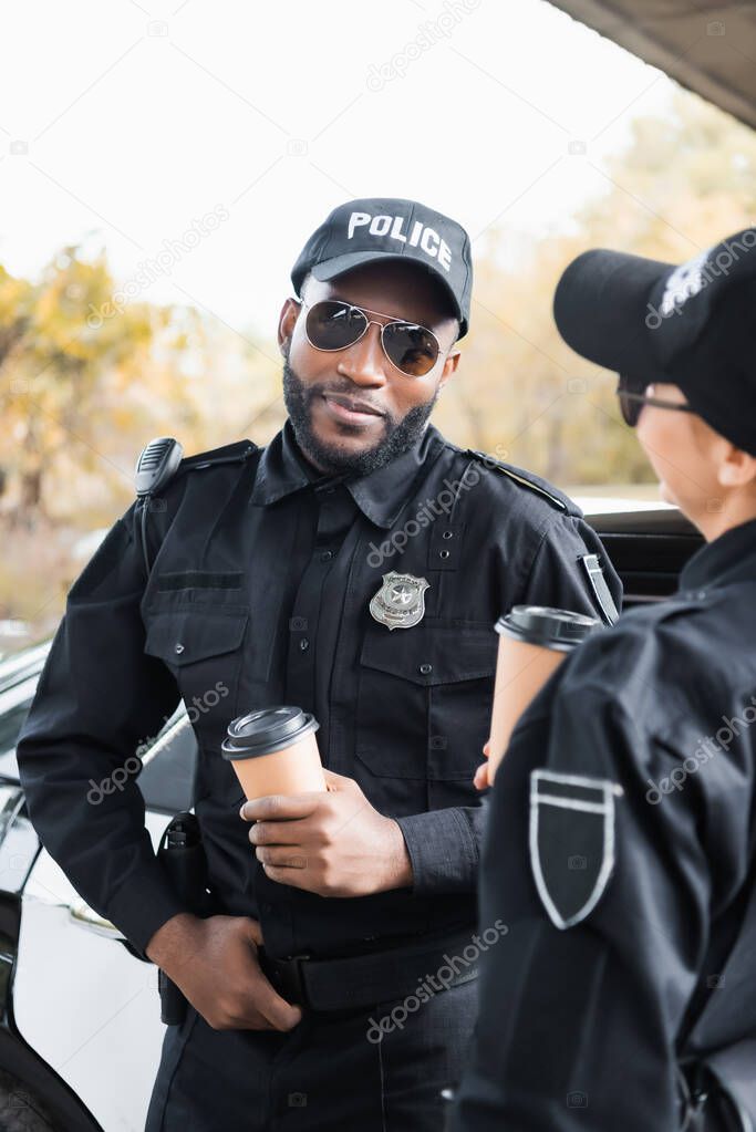 smiling african american police officer with paper cup looking at camera near colleague on blurred background outdoors