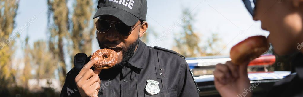 african american police officer eating doughnut with blurred colleague on foreground outdoors, banner