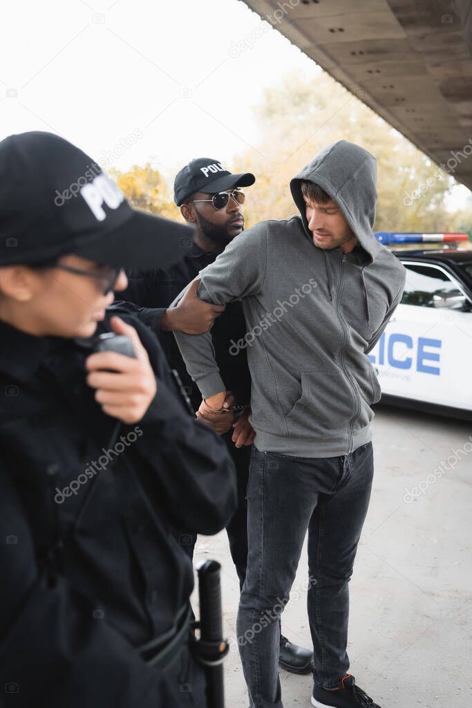 african american policeman handcuffing hooded offender with blurred colleague on foreground outdoors