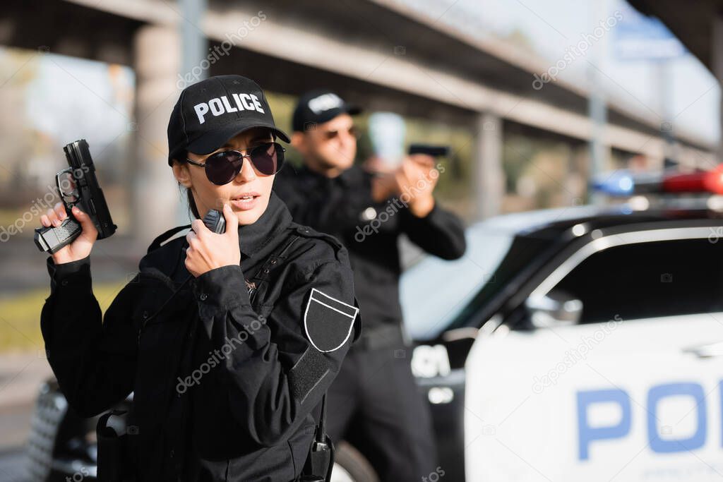 Policewoman in sunglasses holding gun and using walkie talkie near colleague and car on blurred background 