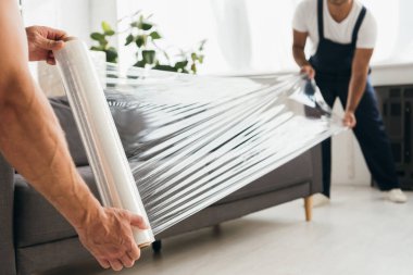 cropped view of movers holding stretch film roll near sofa clipart