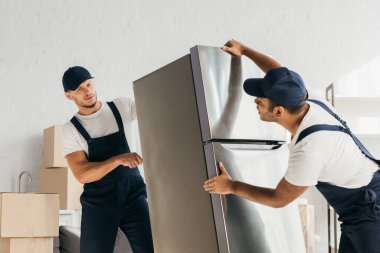 Üniformalı çok kültürlü nakliyeciler dairedeki buzdolabını taşıyorlar. 