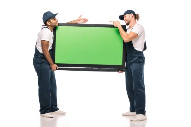 full length of surprised multicultural movers pointing at each other while carrying plasma tv with green screen and on white clipart