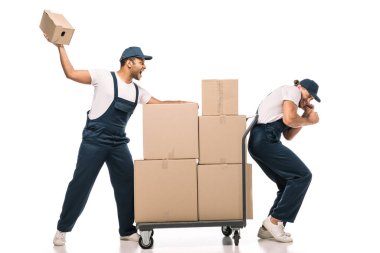 Üniformalı kızgın Hintli nakliyeci elinde karton kutu, korkmuş iş arkadaşının yanında ve beyaz paketli el arabası.