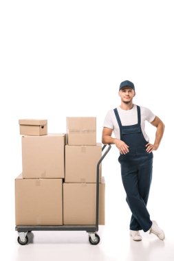 full length of cheerful mover in uniform and cap leaning on hand truck with carton boxes on white clipart