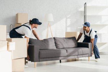 Üniformalı mutlu çok kültürlü nakliyeciler dairede kanepe taşırken gülümsüyorlar.