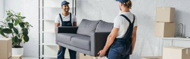 smiling multiethnic movers in uniform carrying couch in apartment, banner clipart