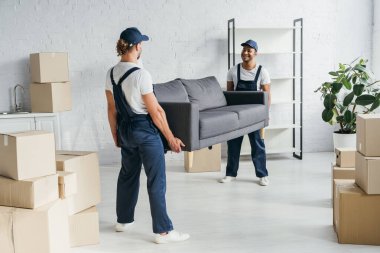 full length of happy young multiethnic movers in uniform carrying couch in apartment clipart
