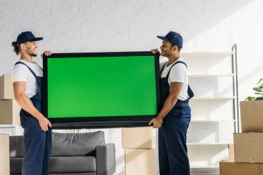 side view of happy multicultural movers in uniform carrying plasma tv with green screen in apartment  clipart