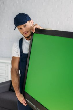 young mover in uniform carrying plasma tv with green screen in apartment  clipart