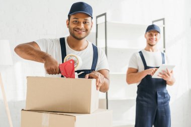 Bulanık arkaplanda dijital tabletle çalışan iş arkadaşının yanında gülümseyen Hintli taşıyıcı paketleme kutusu 