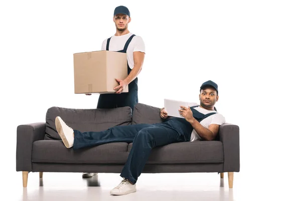 震惊的印地安人拿着数码平板电脑 躺在靠近搬运工的沙发上 盒子在白色上 — 图库照片