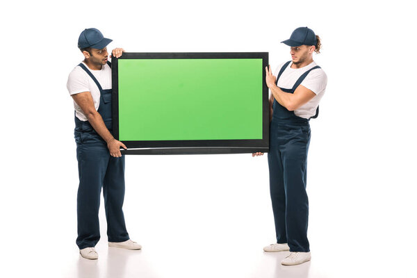 full length of surprised multicultural movers carrying plasma tv with green screen on white