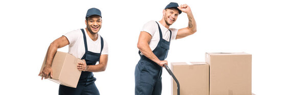 excited multicultural movers in uniform and caps near hand truck with carton boxes on white, banner