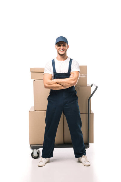 full length of cheerful mover in uniform and cap standing with crossed arms near hand truck with carton boxes on white