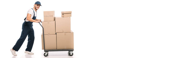 full length of cheerful mover in uniform and cap pulling hand truck with carton boxes on white, banner