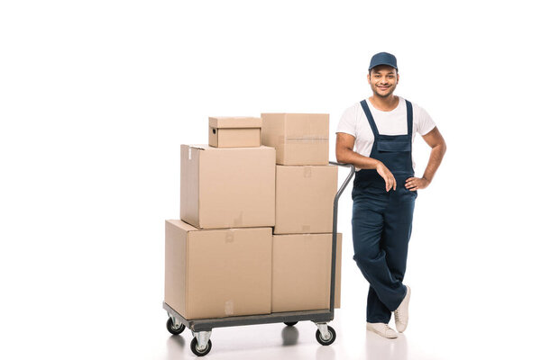 full length of cheerful indian mover in overalls standing with hand on hip near hand truck with carton boxes on white