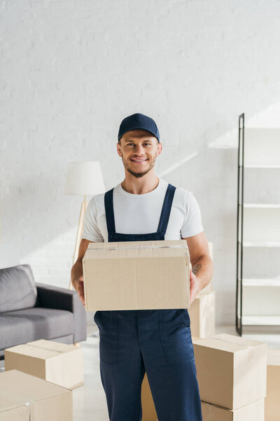 tattooed and smiling mover in cap holding carton box and looking away in apartment 