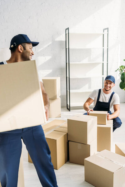 cheerful indian mover in uniform and cap carrying box near happy coworker on blurred background